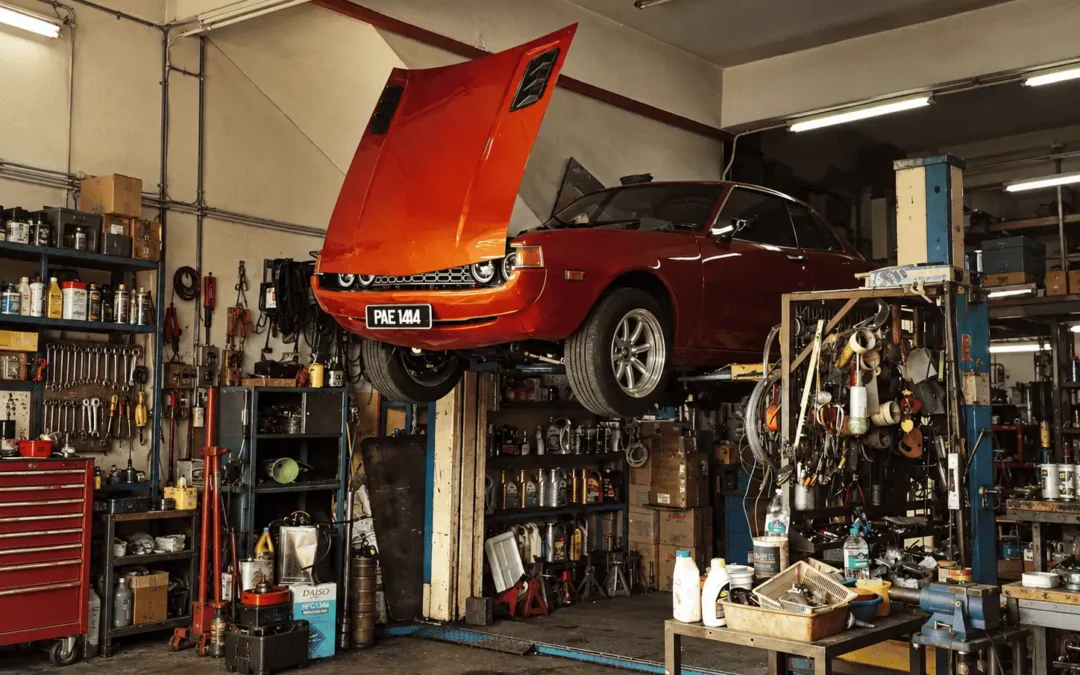 Orange classic car raised on a lift with its hood open, surrounded by tools and shelves in a workshop.